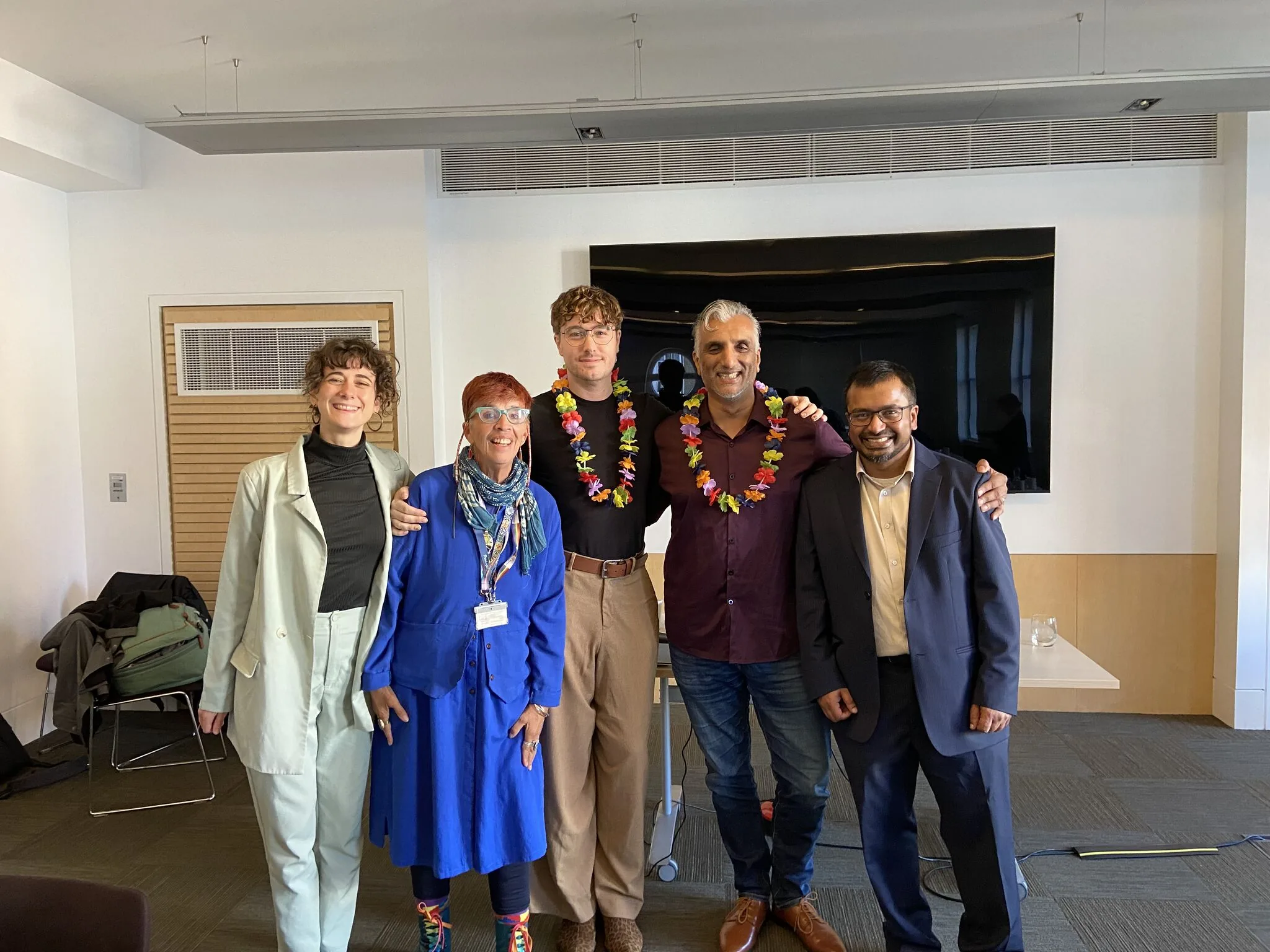 Dr Sarju and a colleague wearing bright flower garlands, standing arm in arm with three other colleagues in a university meeting room with a wall screen behind them.