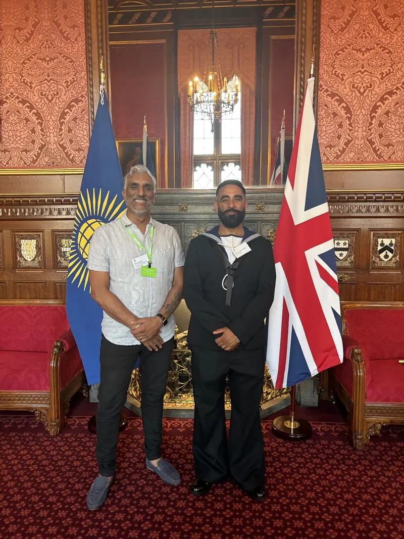 Dr Sarju in a light shirt and lanyard standing beside a person in naval uniform, between the Commonwealth flag and the Union flag in a wood panelled state room hung with red damask.