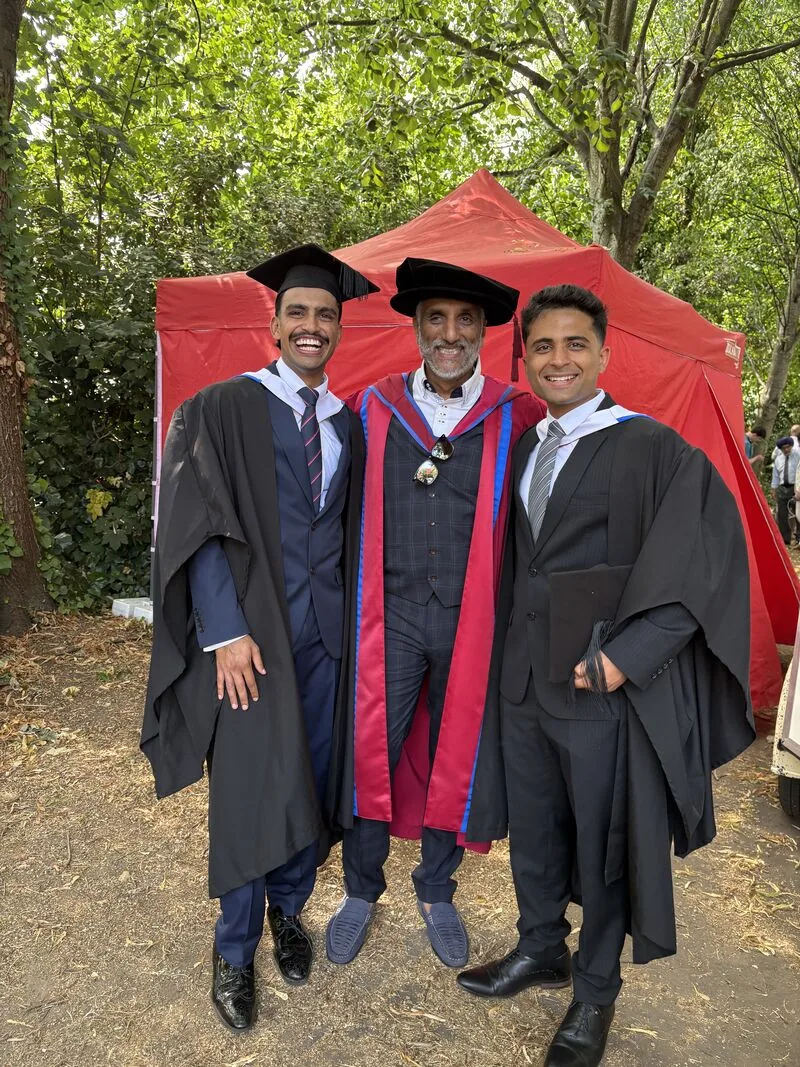 Dr Sarju in doctoral robes and bonnet standing between two graduates in gowns on graduation day.