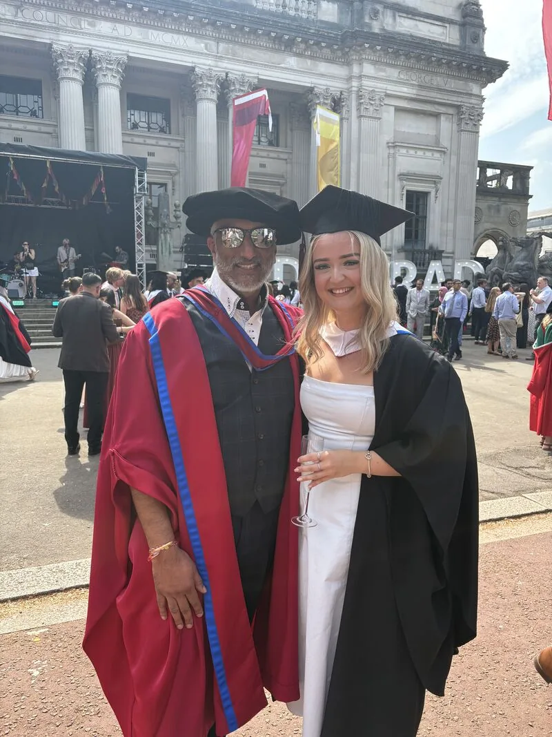 Dr Sarju in red doctoral robes, a bonnet and sunglasses beside a graduate in cap and gown outside a columned university building, with a stage and a crowd behind them.