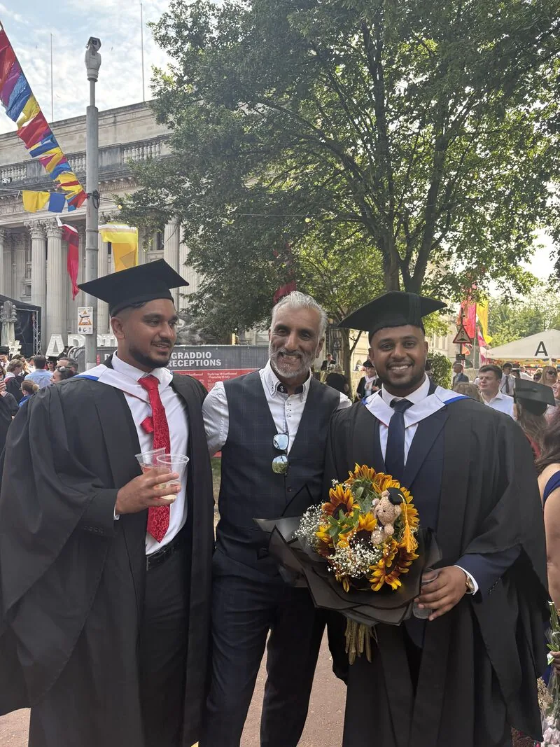 Dr Sarju in a checked waistcoat between two graduates in caps and gowns, one holding a bouquet of sunflowers, at an outdoor graduation reception with bunting overhead.