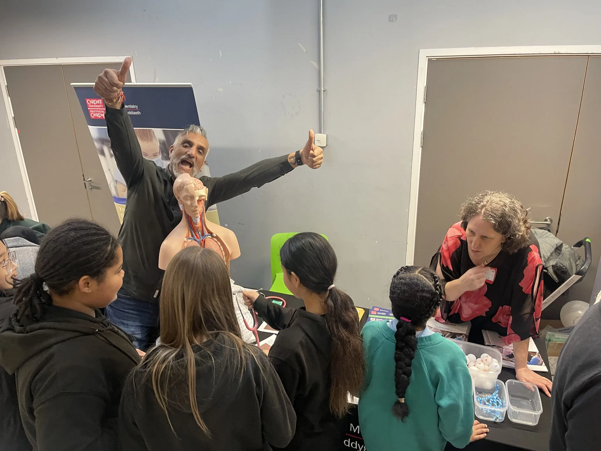 Dr Sarju giving a double thumbs up behind an anatomical torso model while a group of school pupils crowd around the table at a Cardiff University widening access event.