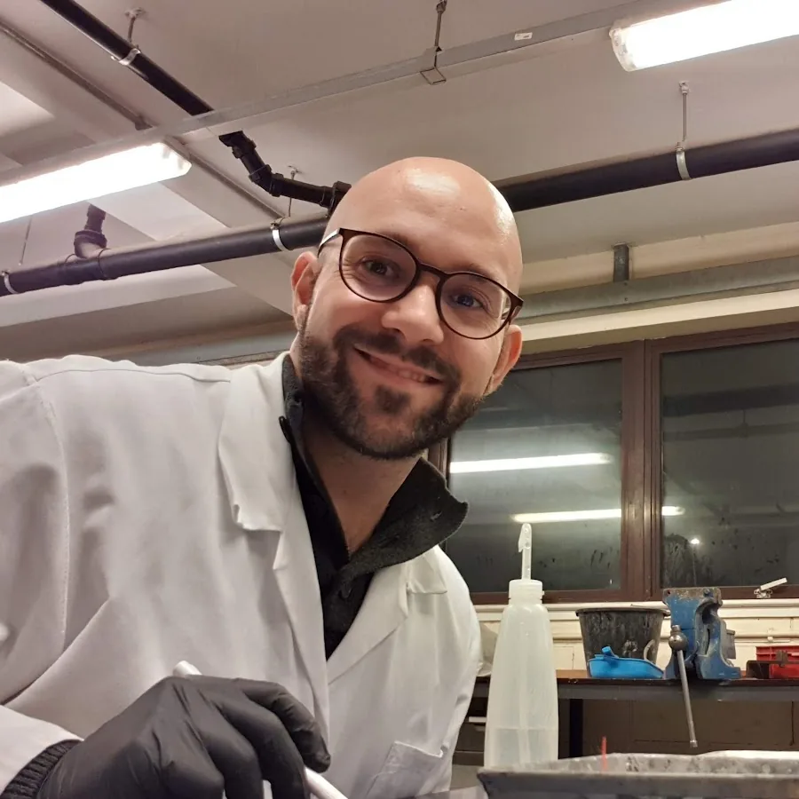 Dr Riccardo Maddalena in a lab coat and black gloves, smiling, at a bench in a materials laboratory.
